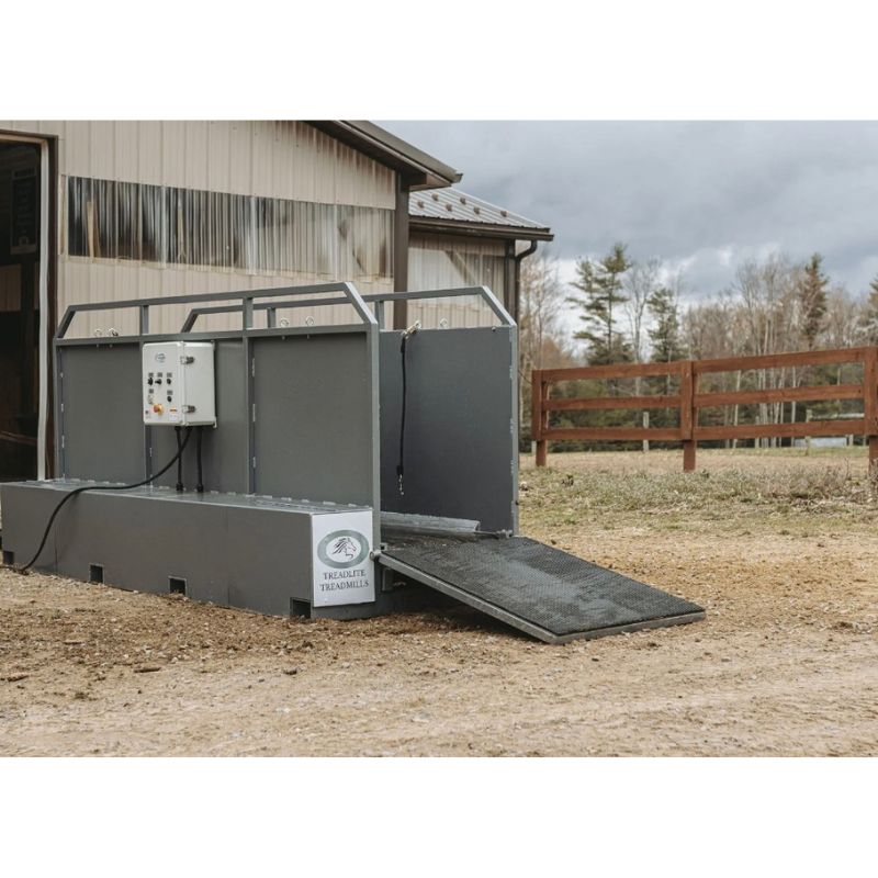 Gray horse treadmill ramp with a control panel in front of a building with a wooden fence and trees in the background.